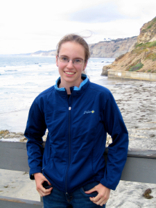 Image is of Elizabeth Sibert, a white woman with glasses and a ponytail wearing a blue zip-up jacket. She is leaning on the railing of Scripps Pier with her back to a beach and ocean in the background.